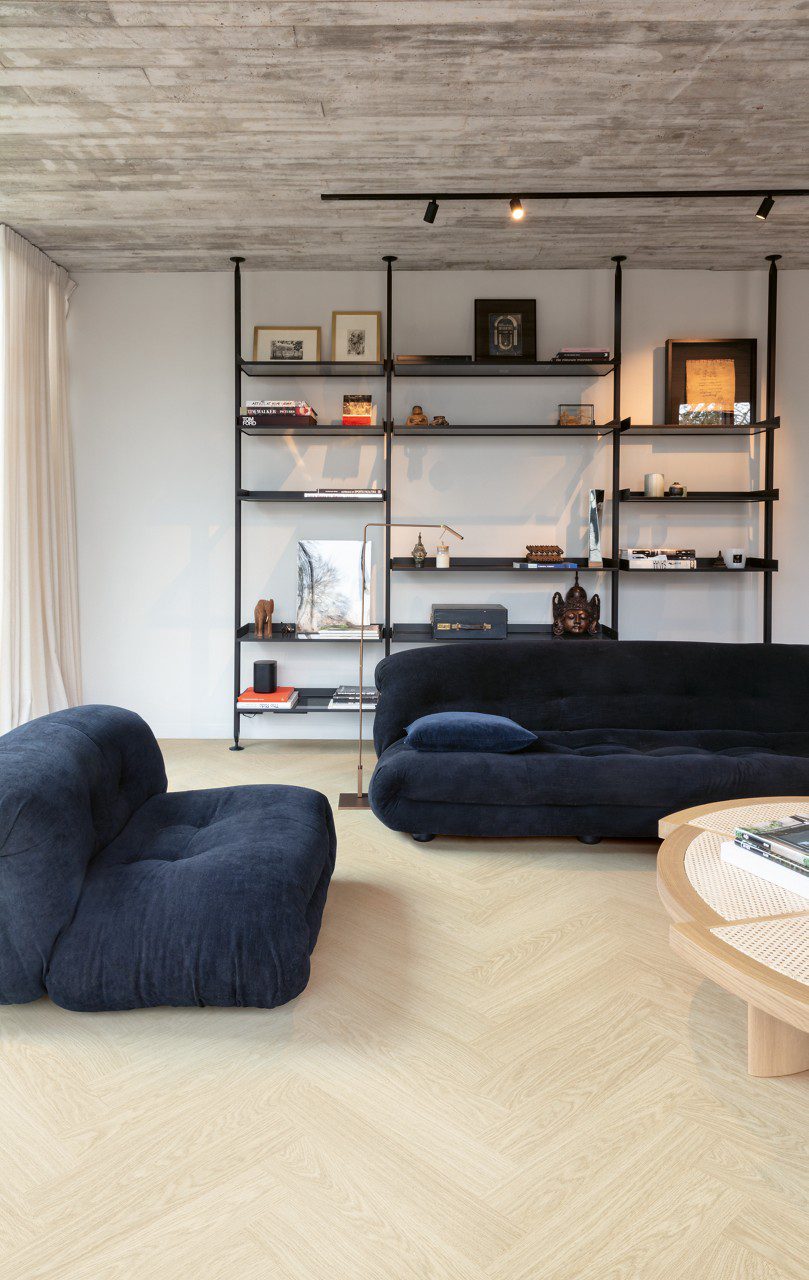 Vinyl Fischgrat Quick Step Ciro Pure Oak Polar, verlegt im Wohnzimmerboden, mit blauem Sofa, Couchtisch und Bücherregal.