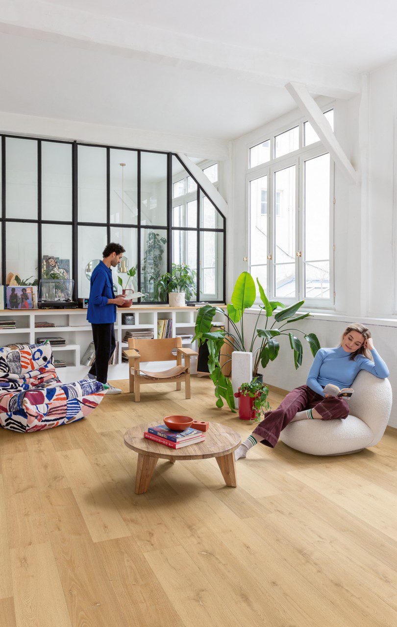 LVT flooring Quick-Step Bloom Elegant Oak Natural installed on livingroom floor. Man is looking a vinyl record and woman is sitting in a chair and reading a book.