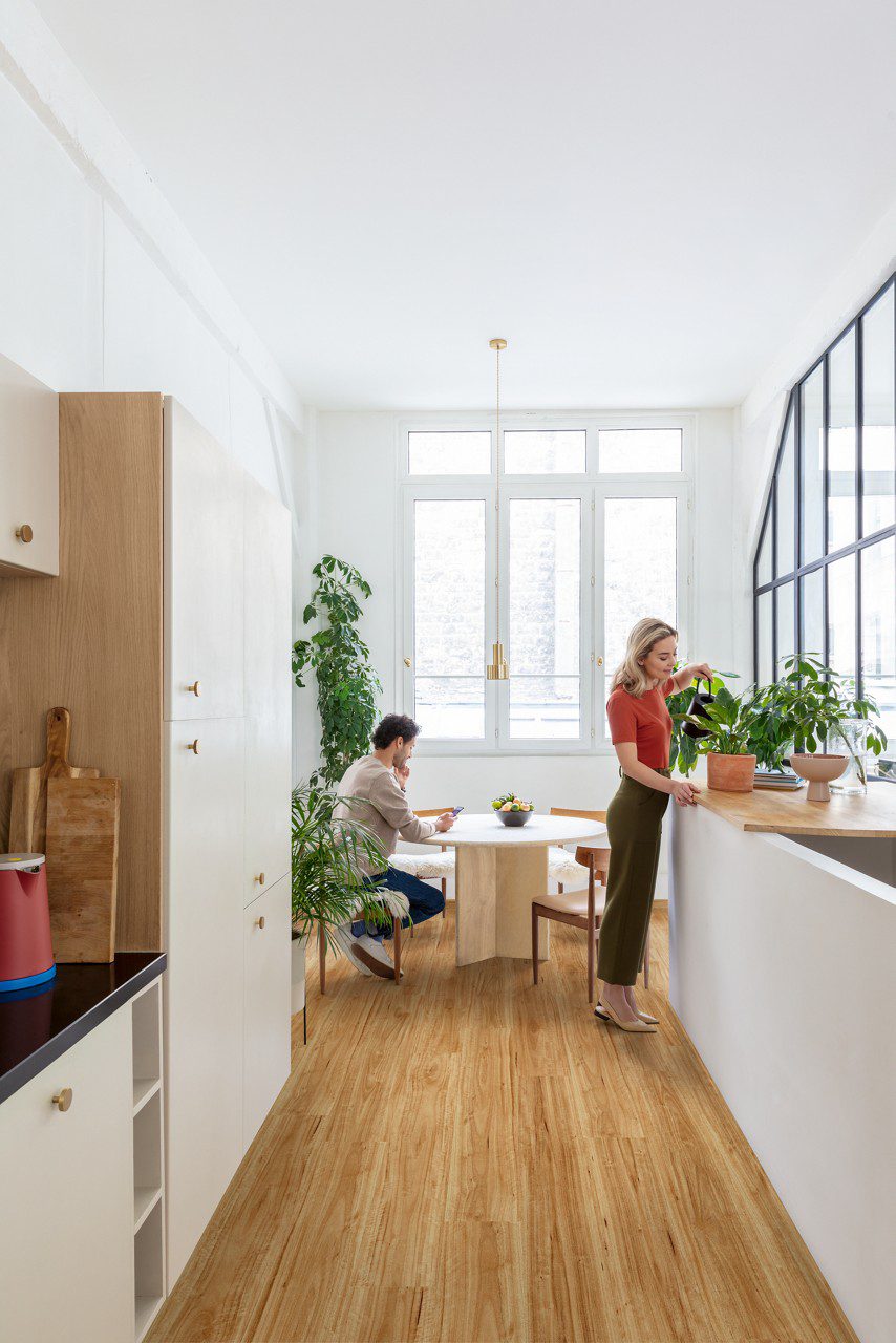 Vinyylilankku Quick Step Bloom Urban Blackbutt asennettuna keittiön lattialle, nainen kastelee keittiötasolla olevaa ruukkukasvia, mies istuu ruokapöydän ääressä ja katsoo puhelinta.