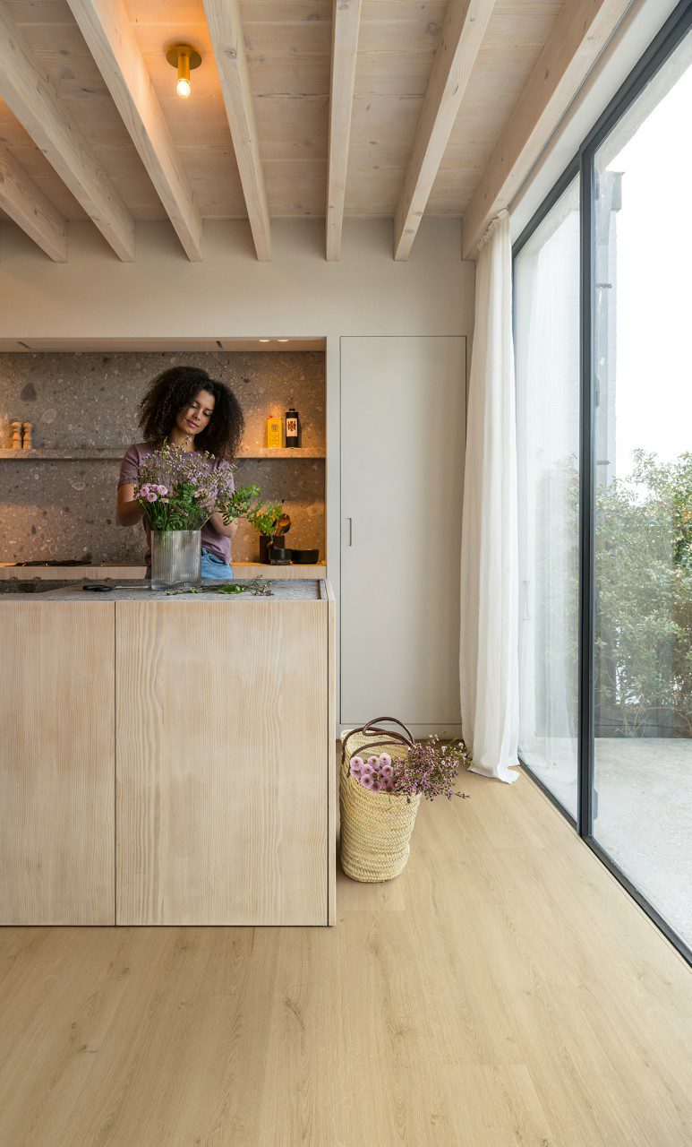 Vinyylilattia Quick Step Bloom Botanic Beige asennettuna keittiön lattialle, nainen kohentaa keittiösaarella olevaa ruukkukasvia, vaalea luonnonbeige sävy ja lankkudesign.