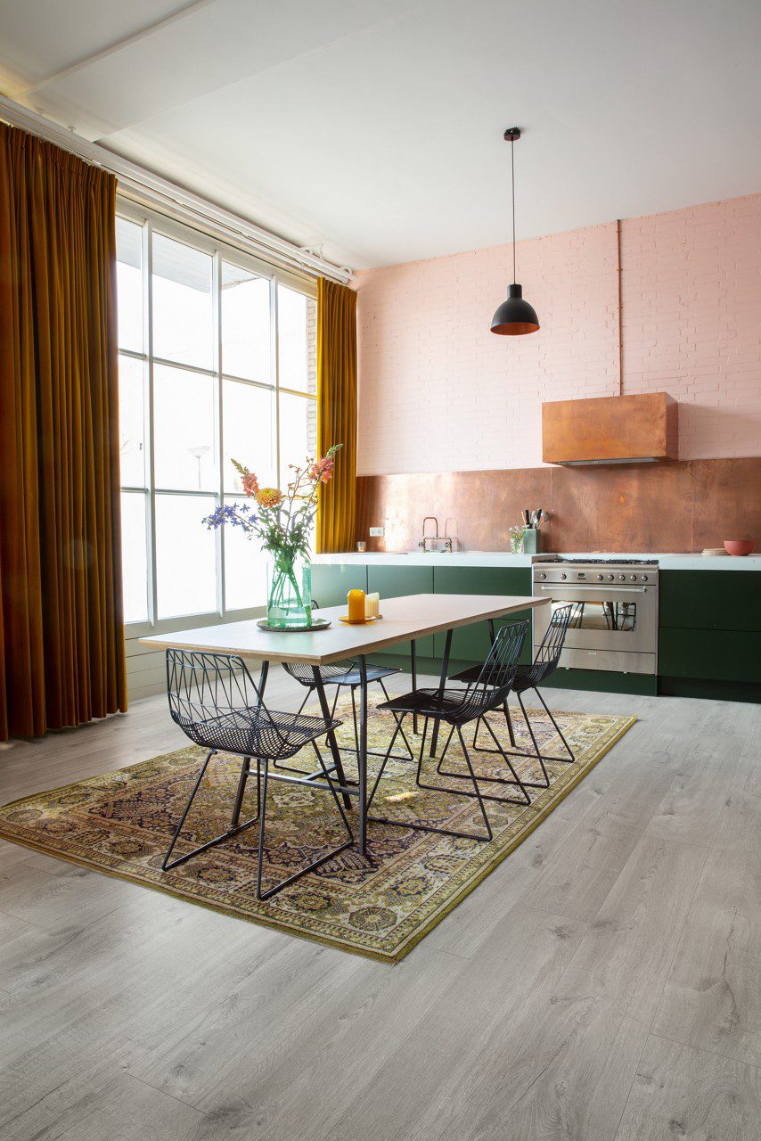 Vinyylilattia Quick Step Bloom Cotton Oak Cold Grey asennettuna keittiön lattialle, viileä vaaleanharmaa tammen sävy ja lankkudesign, keittiökalusteet tyylikkäällä kuparinsävyisellä taustalevyllä, ruokapöytä, tuolit ja kukkia pöydällä.