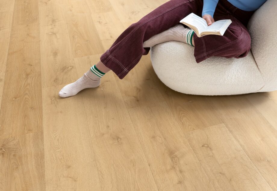 виниловыи-ламинат-quick-step-bloom-elegant-oak-natural Виниловый ламинат Quick Step Bloom Elegant Oak Natural уложен на полу в гостиной, женщина сидит в удобном кресле и читает книгу.