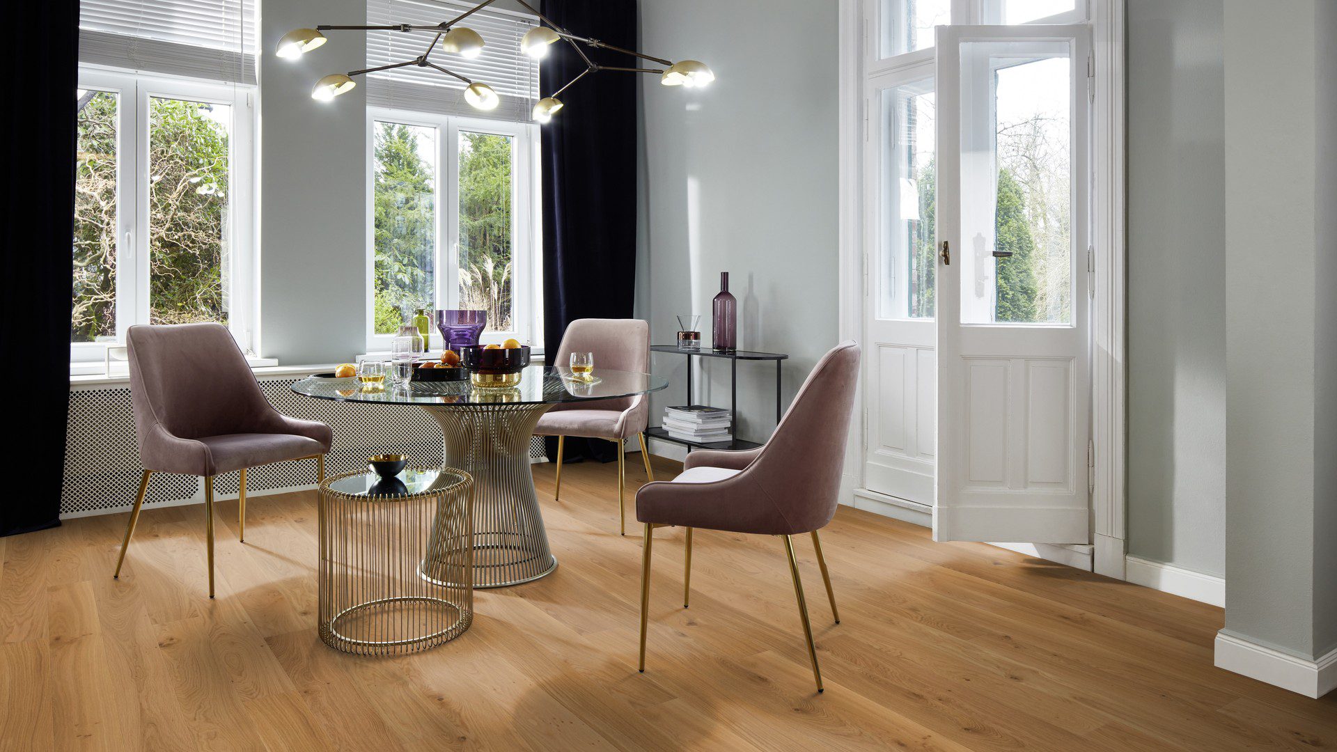 Lankkuparketti Bauwerk Boen Casapark Budget Cut tammi 35, harjattu ja mattalakattu, asennettuna ruokailutilan lattialle – tyylikäs lasikantinen ruokapöytä hedelmineen ja juomalaseineen, lilansävyiset pehmeät tuolit sekä avoin parvekkeen ovi. Luonnollinen, lämminsävyinen tammensävy ja kevyesti rustiikkinen sorteerus (maltillisesti oksakohtia ja hienovaraista sävyvaihtelua).