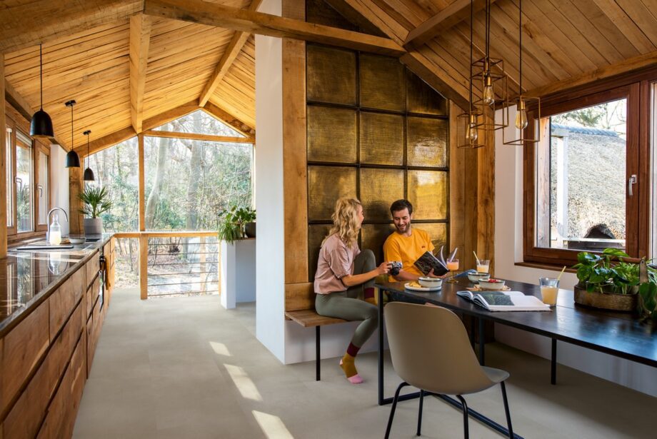 LVT põrandakate Quick-Step Illume Sandstone concrete helehalli beezhika tooniga paigaldatud küügi põrandale. rustikaalne stiil toetavatest puidust prussidest ja laudadest laega. Köögimööbel, Mees istub laua taga avatud ajakiri käes ja naine näitab fotoaparaadist pilte.