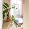 klick-vinylboden-quick-step-bloom-brushed-oak-beige Klick Vinylboden Quick Step Bloom Brushed Oak Beige im Schlafzimmer verlegt, mit Palme, rosa Sitzhocker, rosa Vorhängen und Bett.