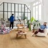lvt-floors-quick-step-bloom-elegant-oak-natural LVT floors Quick-Step Bloom Elegant Oak Natural installed on a living room floor. Woman is sitting on a chair and reading a book. Man is looking the vinyl record player. Table with books, palm threes