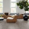 LVT parkett Quick-Step Bloom Cotton Oak Cozy Grey paigaldatud elutoa põrandale. Pruunist nahast dugitool, diivanilaud lilledega, palmipuu, pikk kummut akende all.