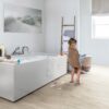 veekindel-vinuulparkett-quick-step-blos-canyon-tamm-beige Veekindel vinüülparkett Quick-Step Blos Canyon tamm beige paigaldatud vannitoa põrandale. Valge kandiline vann on täis vannivahtu ja väike tüdruk on vannist välja tulnud ning on käterätis. Põrand on koos vannivahuse veega. Kuivaturedel millel on särk kuivamas ja seinal on pilt liivasest rannast koos kasvavate heintega.