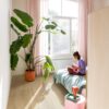 vinyl-floor-quick-step-bloom-brushed-oak-beige Quick Step Bloom Brushed Oak Beige vinyl floor installed in a bedroom, with a palm plant, pink curtains, and a woman reading a book on the bed.