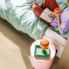 vinylboden-quick-step-bloom-brushed-oak-beige Vinylboden Quick Step Bloom Brushed Oak Beige im Schlafzimmer, Frau liest ein Buch auf dem Bett, rosa Sitzhocker.