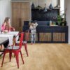 vinyylilattia-quick-step-blos-canyon-tammi-natural Vinyylilattia Quick Step Blos Canyon Tammi Natural asennettuna ruokailutilan lattialle, luonnollinen tammen sävy ja lankkulattia tammi, äiti istuu ruokapöydän ääressä ja seuraa tytärtä, joka ottaa banaanin hedelmävadilta keittiön kaapista.