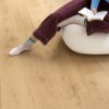 виниловыи-ламинат-quick-step-bloom-elegant-oak-natural Виниловый ламинат Quick Step Bloom Elegant Oak Natural уложен на полу в гостиной, женщина сидит в удобном кресле и читает книгу.