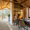 LVT põrandakate Quick-Step Illume Sandstone concrete helehalli beezhika tooniga paigaldatud küügi põrandale. rustikaalne stiil toetavatest puidust prussidest ja laudadest laega. Köögimööbel, Mees istub laua taga avatud ajakiri käes ja naine näitab fotoaparaadist pilte.