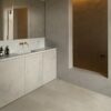 Vinylboden in Betonoptik Quick-Step Illume Sandstone Concrete, verlegt auf dem Badezimmerboden – graues Badmöbel mit grauer Waschtischplatte und Aufsatzbecken, drei Spiegelschränke an der Wand sowie eine goldene Wandarmatur; warmer Greige-/Sandton mit dezentem Betondesign.