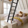 Кварц-винил Quick-Step Illume Sandstone Concrete уложен на полу мансардной спальни — женщина лежит в постели и читает книгу; тёплый песочно-бежевый (greige) оттенок и спокойный бетонный дизайн.